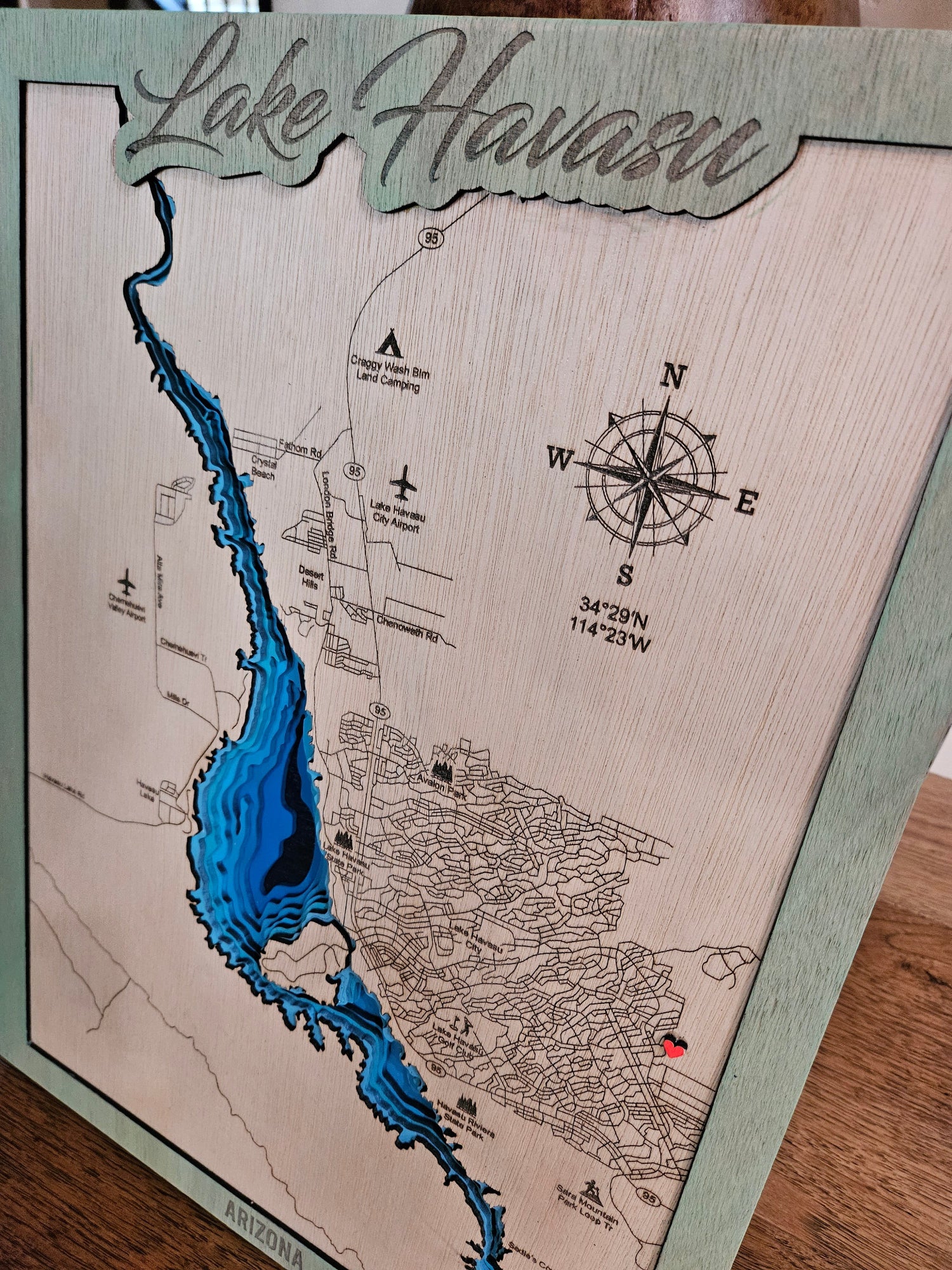 A 3D laser-cut wooden Lake Havasu map plaque with a blue depth-effect design, a compass rose, and a green border, shown on a wooden tabletop.