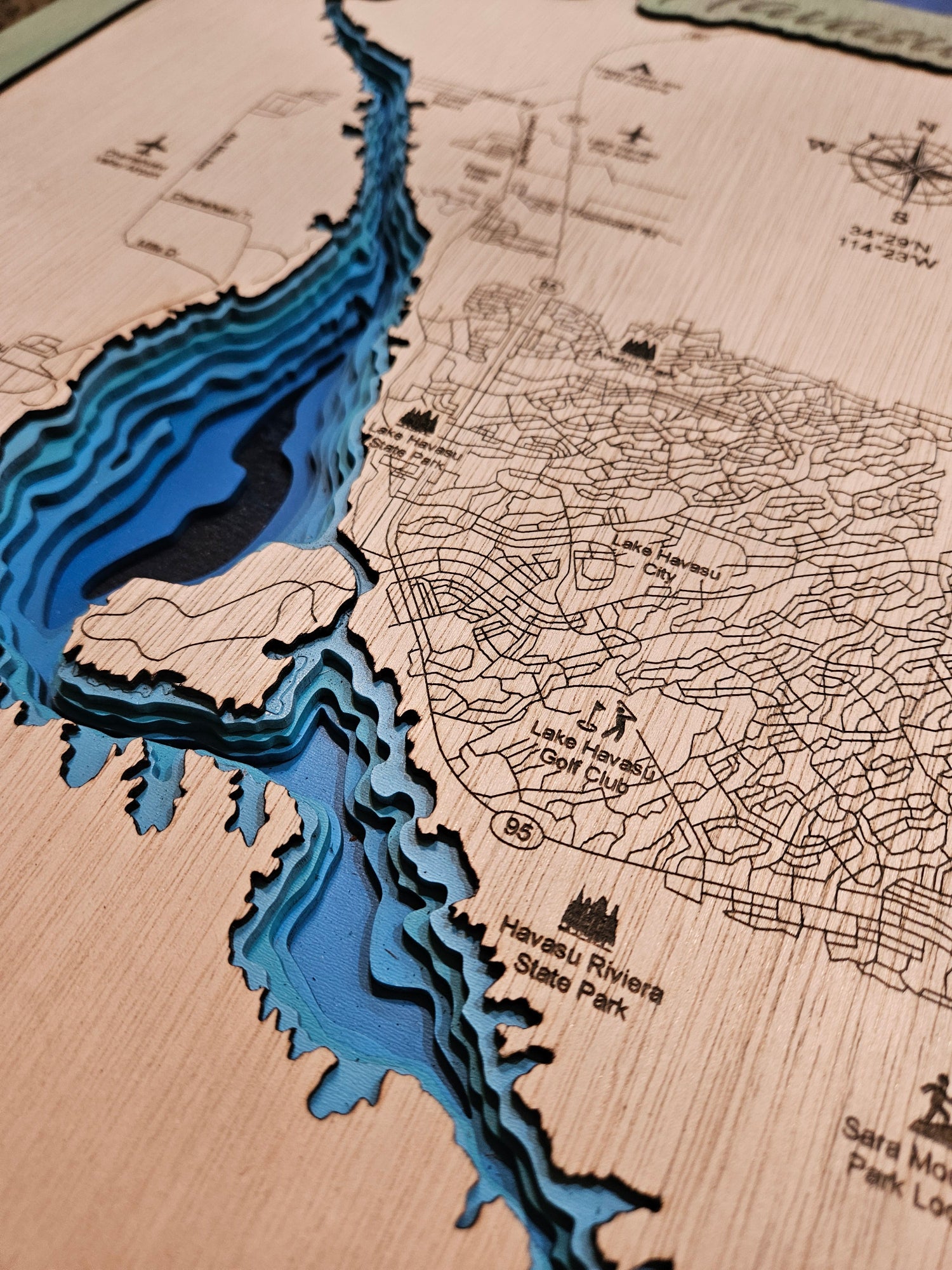 A close-up of a 3D layered wooden map of Lake Tahoe, featuring a prominent blue and black layered river basin and black engraved street and landmark labels on a light wood background.
