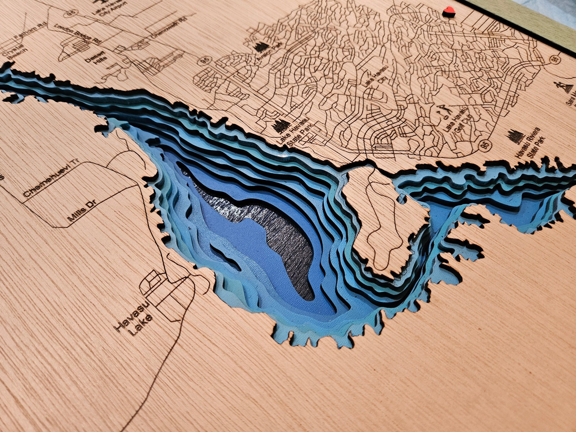 A close-up of a 3D laser-etched wooden topographic map of Hawesee Lake, showing layered wood cutouts that create a blue and black depth effect, with engraved street and lake names on a light wood grain background.