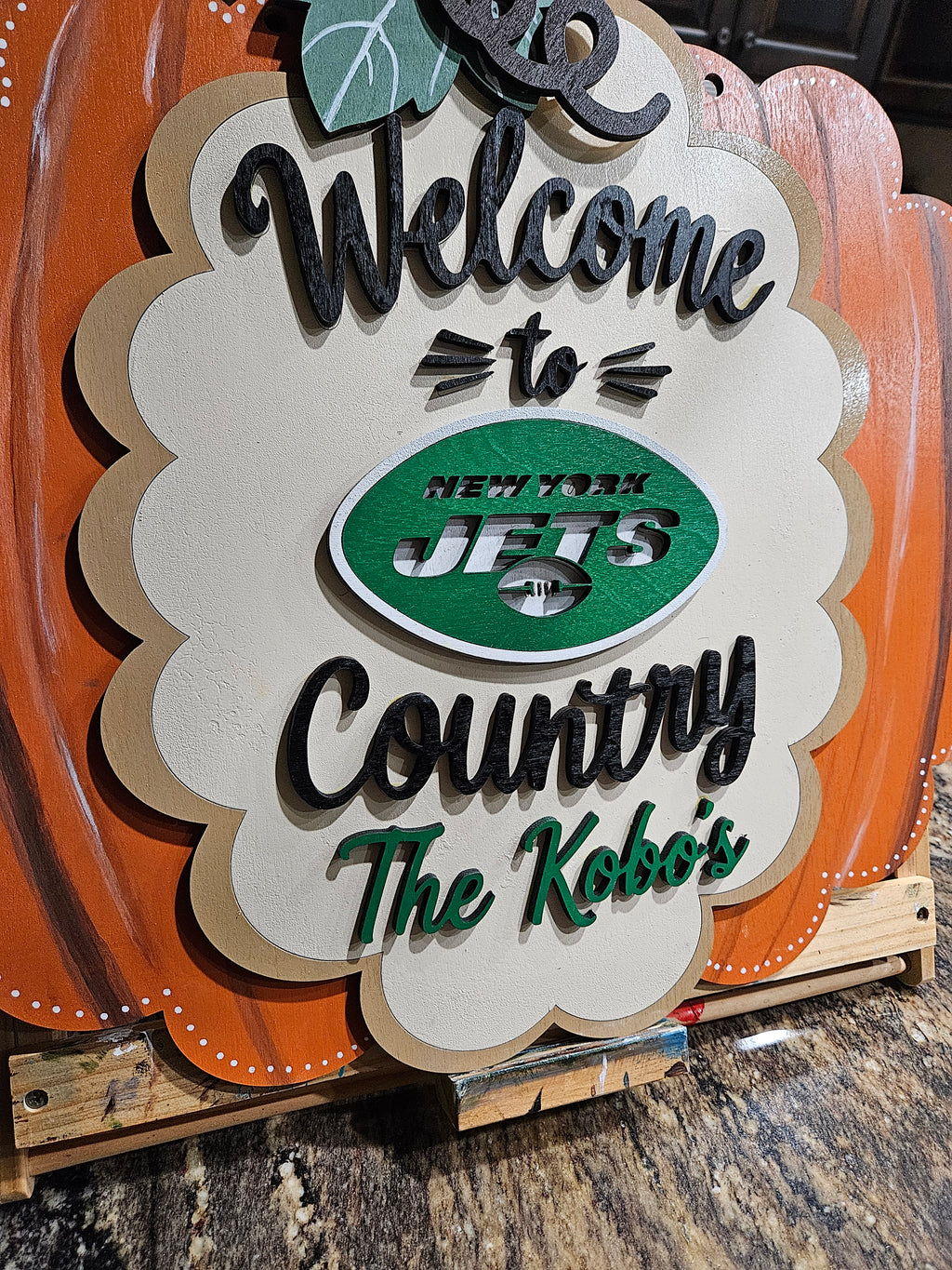 A decorative wooden sign with a scalloped cream center panel and orange pumpkin-like side panels. The sign reads “Welcome to Country The Kobo’s” in black and green lettering, with a green oval New York Jets logo in the middle.