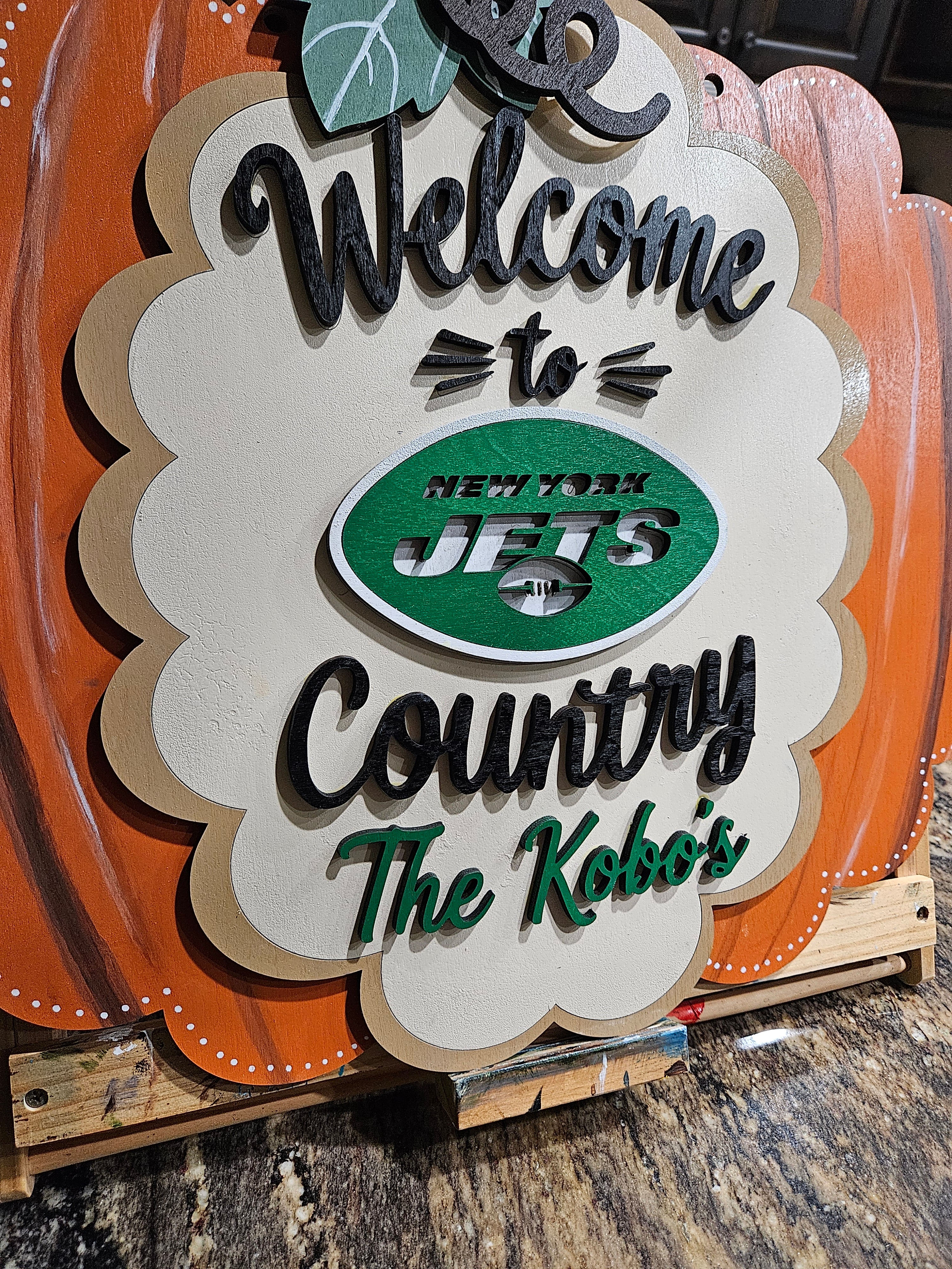A decorative wooden sign with a scalloped cream center panel and orange pumpkin-like side panels. The sign reads “Welcome to Country The Kobo’s” in black and green lettering, with a green oval New York Jets logo in the middle.