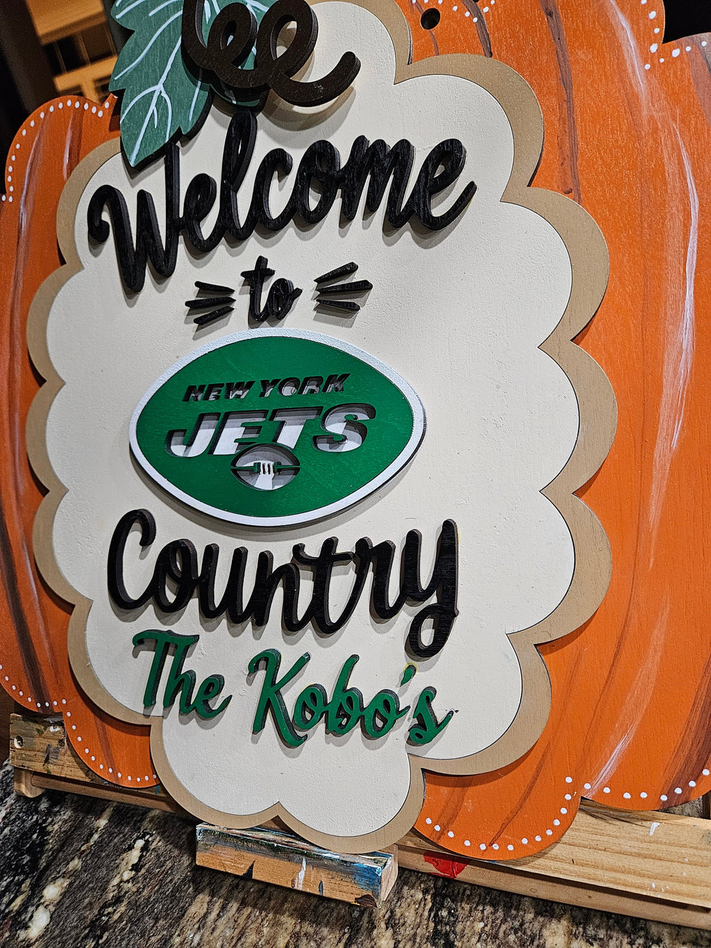 A decorative wooden sign shaped like a pumpkin with a scalloped white center panel reading 'Welcome to Country The Kobo's' and featuring a green oval New York Jets logo. The sign is photographed on a wooden stand over a dark stone surface.