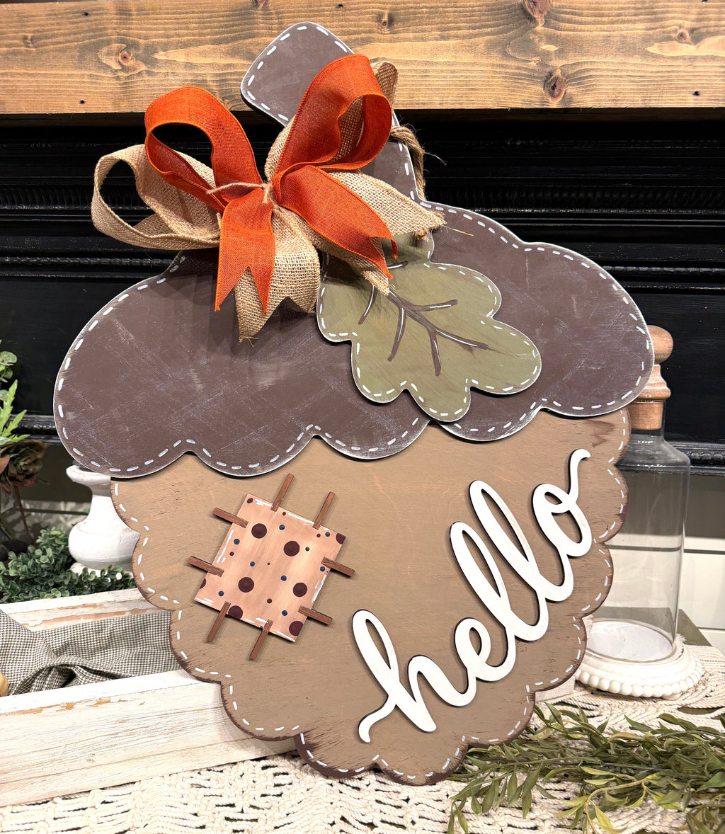 Acorn-shaped decorative wall sign with a brown and tan scalloped edge, a large orange and beige ribbon bow, a green leaf, a small stitched square motif, and the word "hello" in white script, displayed on a tabletop with fall decor.