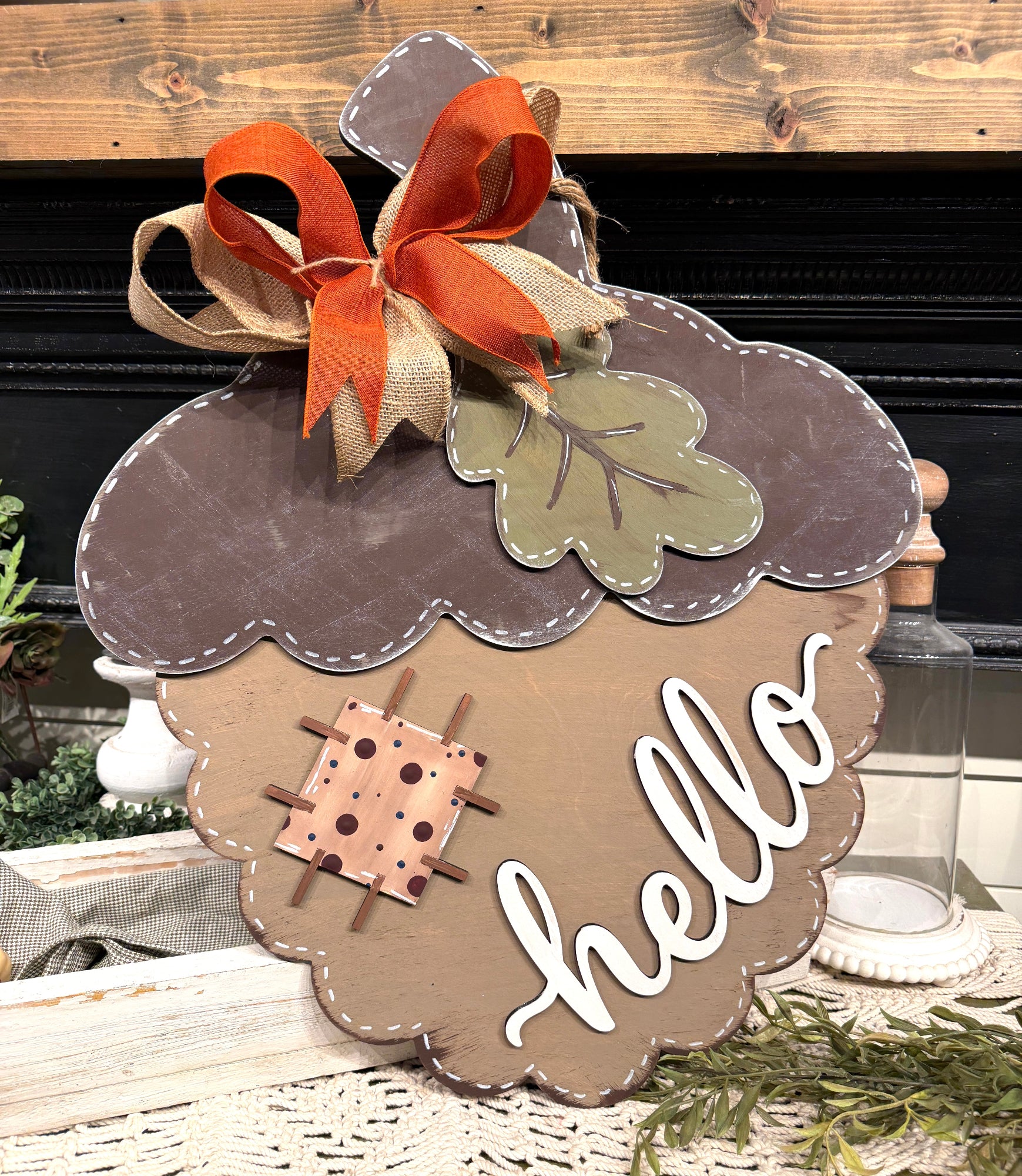 Acorn-shaped decorative wall sign with a brown and tan scalloped edge, a large orange and beige ribbon bow, a green leaf, a small stitched square motif, and the word "hello" in white script, displayed on a tabletop with fall decor.