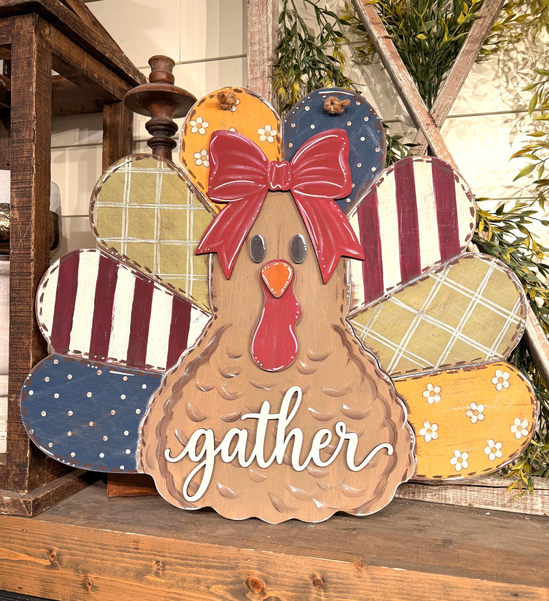 A rustic wooden decorative sign shaped like a turkey, with patterned feathers in red, blue, yellow, and beige, a red bow, and the word "gather" in white script on the front, displayed on a wooden shelf.