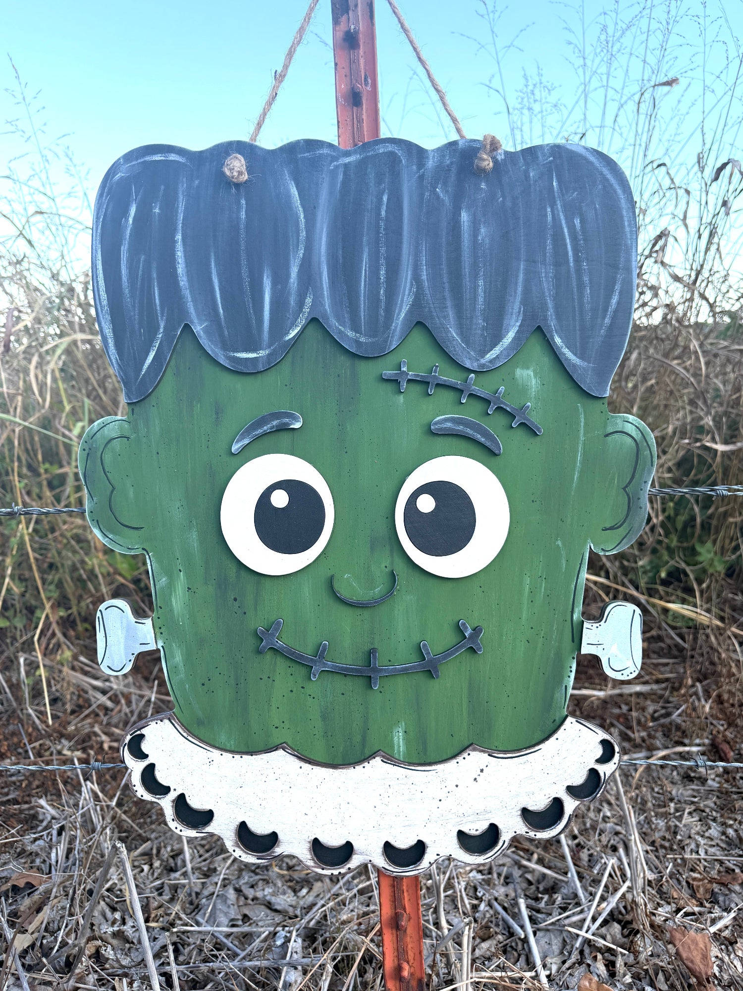 A painted wooden Frankenstein sign hanging from a string outdoors. The sign has green skin, dark blue hair, a white ruffled collar, and stitched facial details, with dry grass and blue sky in the background.