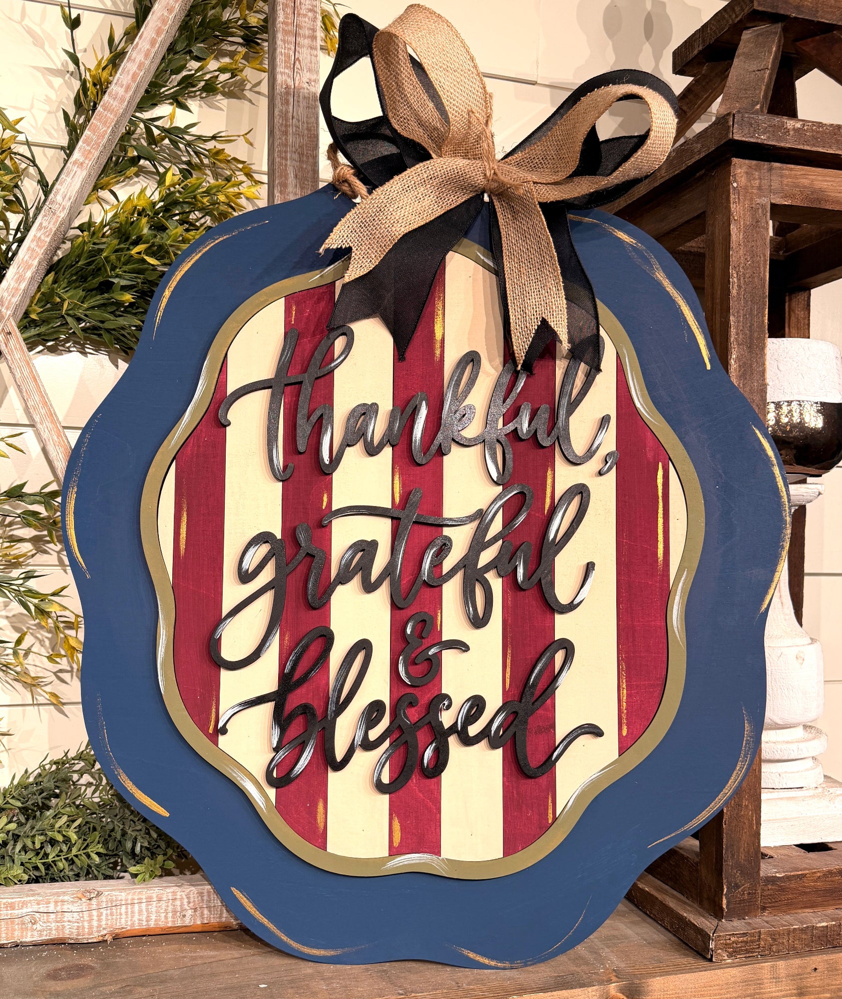 A scalloped autumn-themed wooden sign with a blue border, red and cream vertical stripes, and raised black lettering that reads 'thankful, grateful & blessed.' It has a tan burlap ribbon and black bow hanging from the top, shown in a styled home decor setting.
