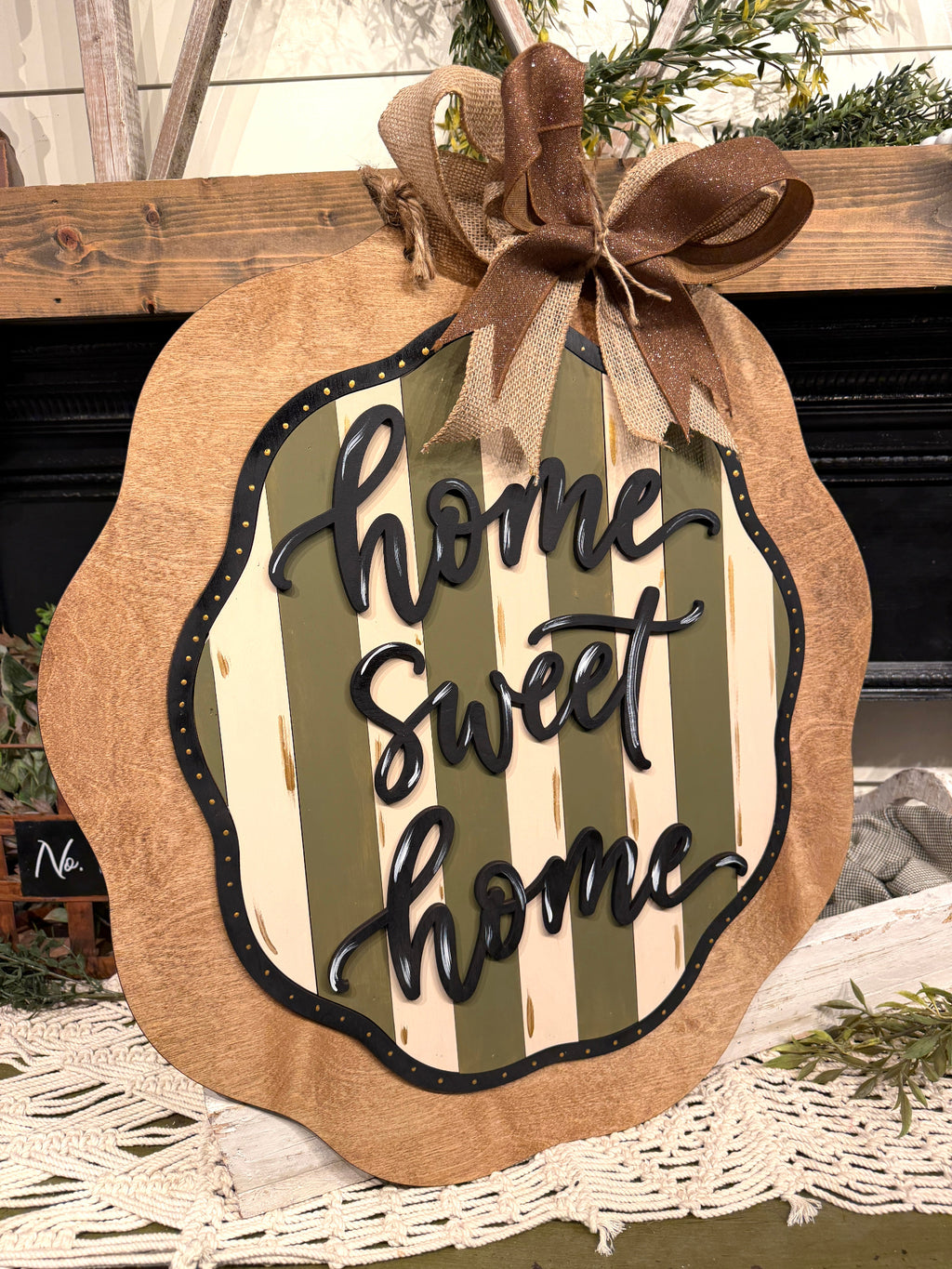 A decorative autumn sign shaped like a pumpkin with a scalloped burlap edge, a brown ribbon bow at the top, and the phrase "home sweet home" in black script over cream and olive green vertical stripes. The sign is displayed in a home setting on a white lace runner in front of a dark fireplace.