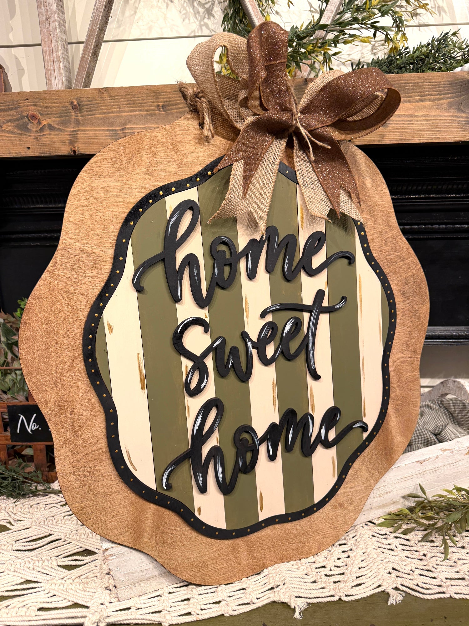 A decorative autumn sign shaped like a pumpkin with a scalloped burlap edge, a brown ribbon bow at the top, and the phrase "home sweet home" in black script over cream and olive green vertical stripes. The sign is displayed in a home setting on a white lace runner in front of a dark fireplace.