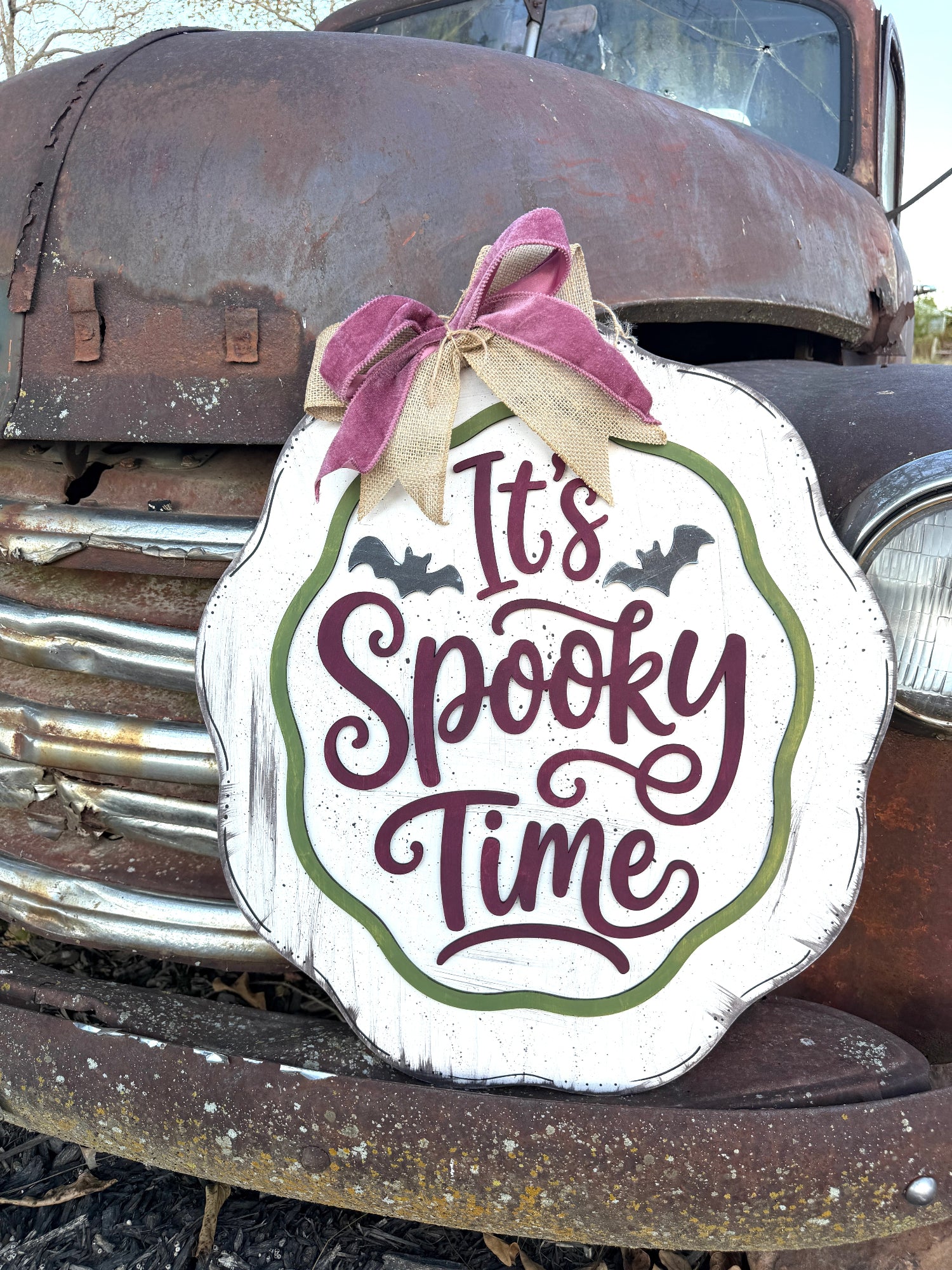 A scalloped white wooden sign with 'It's Spooky Time' in dark purple script, decorated with a purple and tan ribbon bow, displayed against the rusty front of an old truck.