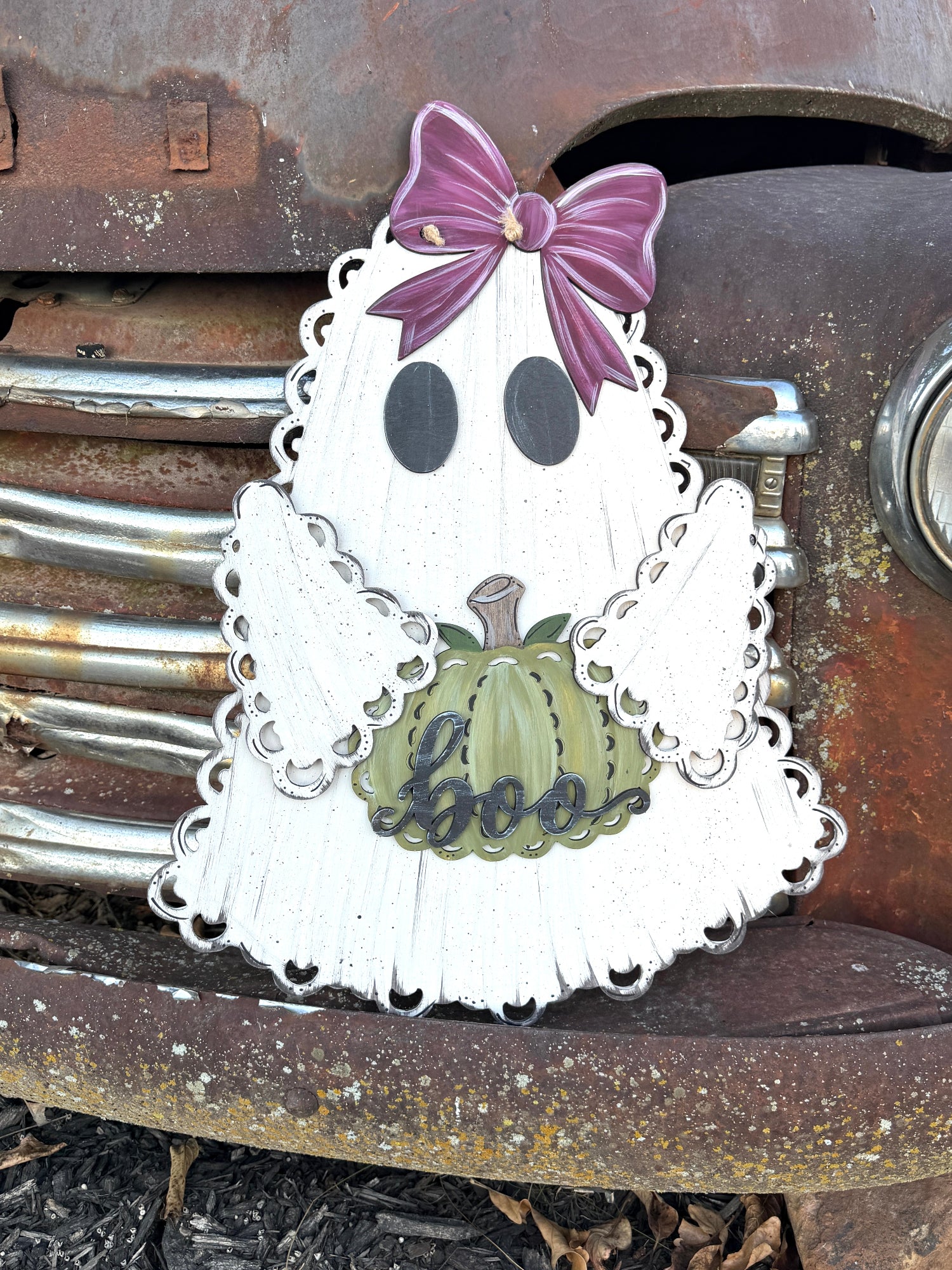A decorative ghost-shaped Halloween sign with a distressed white finish, scalloped edges, black eyes, a purple bow, and a green pumpkin reading 'boo', photographed against a rusty metal surface.