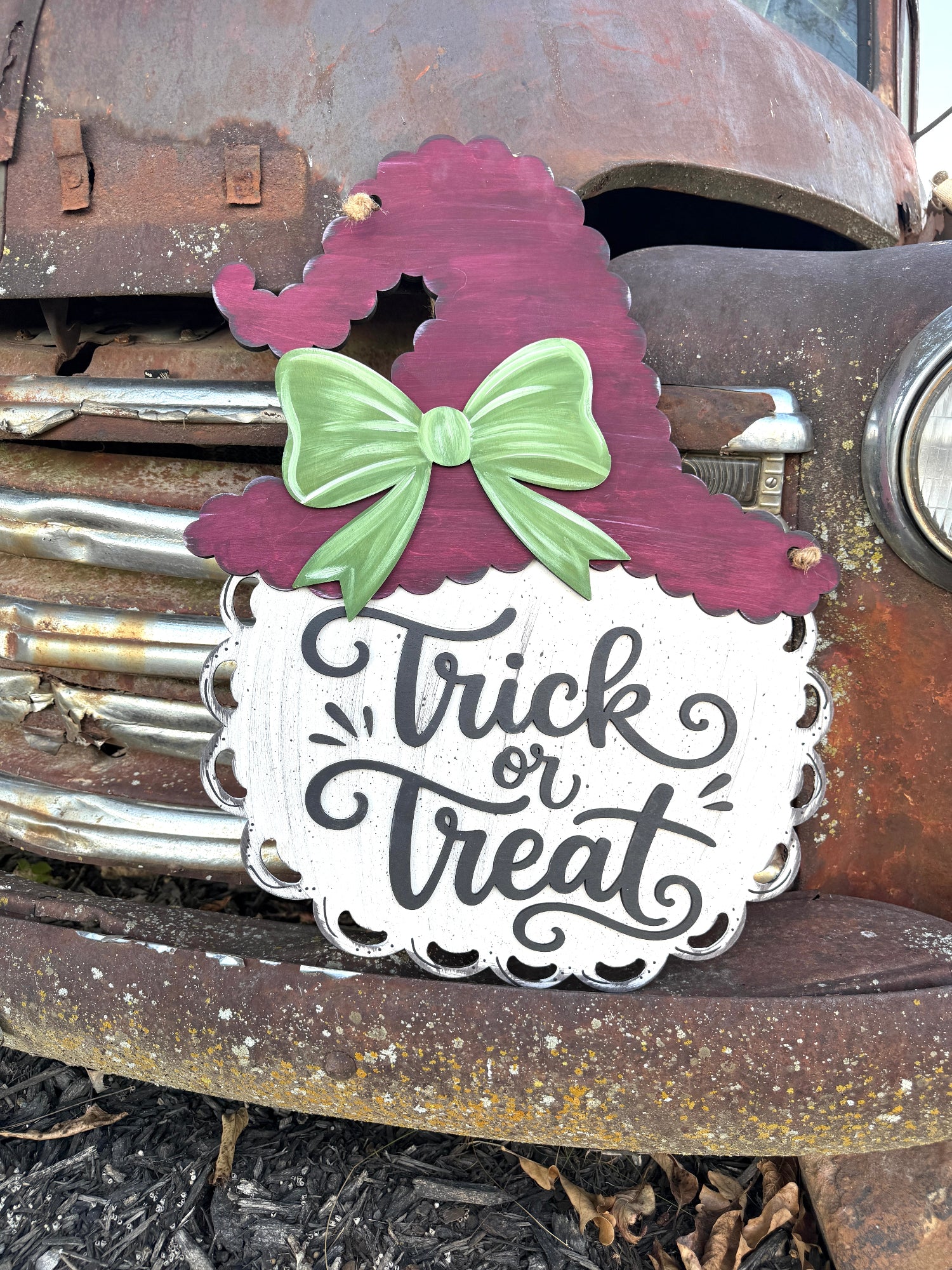 A decorative Halloween sign with a white scalloped base, black script text reading 'Trick or Treat,' a dark burgundy witch hat silhouette, and a light green bow, displayed against a rusted vintage car.