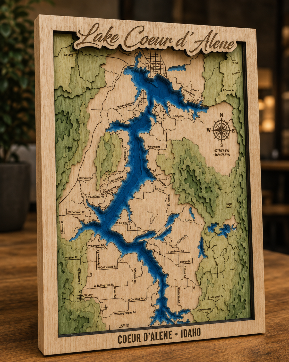 A framed 3D laser-cut wooden map of Lake Coeur d'Alene, Idaho, featuring green topographic terrain, blue water, and engraved text on a wooden tabletop with a blurred indoor background.