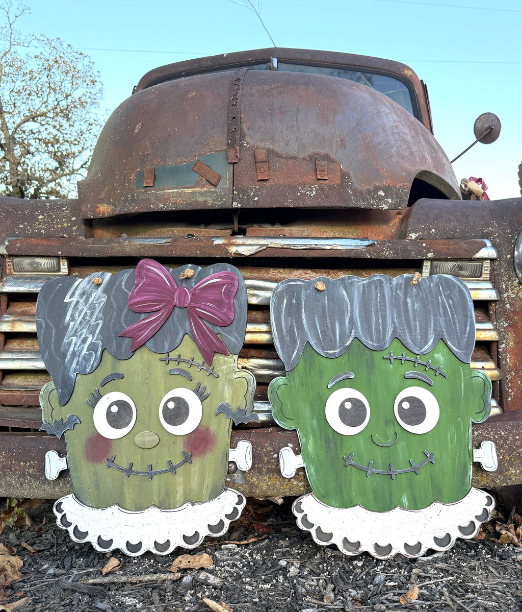 Two painted wooden Frankenstein-themed yard signs in front of a rusted vintage truck outdoors. One sign depicts a green female monster with a purple bow and white ruffled collar, and the other depicts a green male monster with stitched hair and a white ruffled collar.
