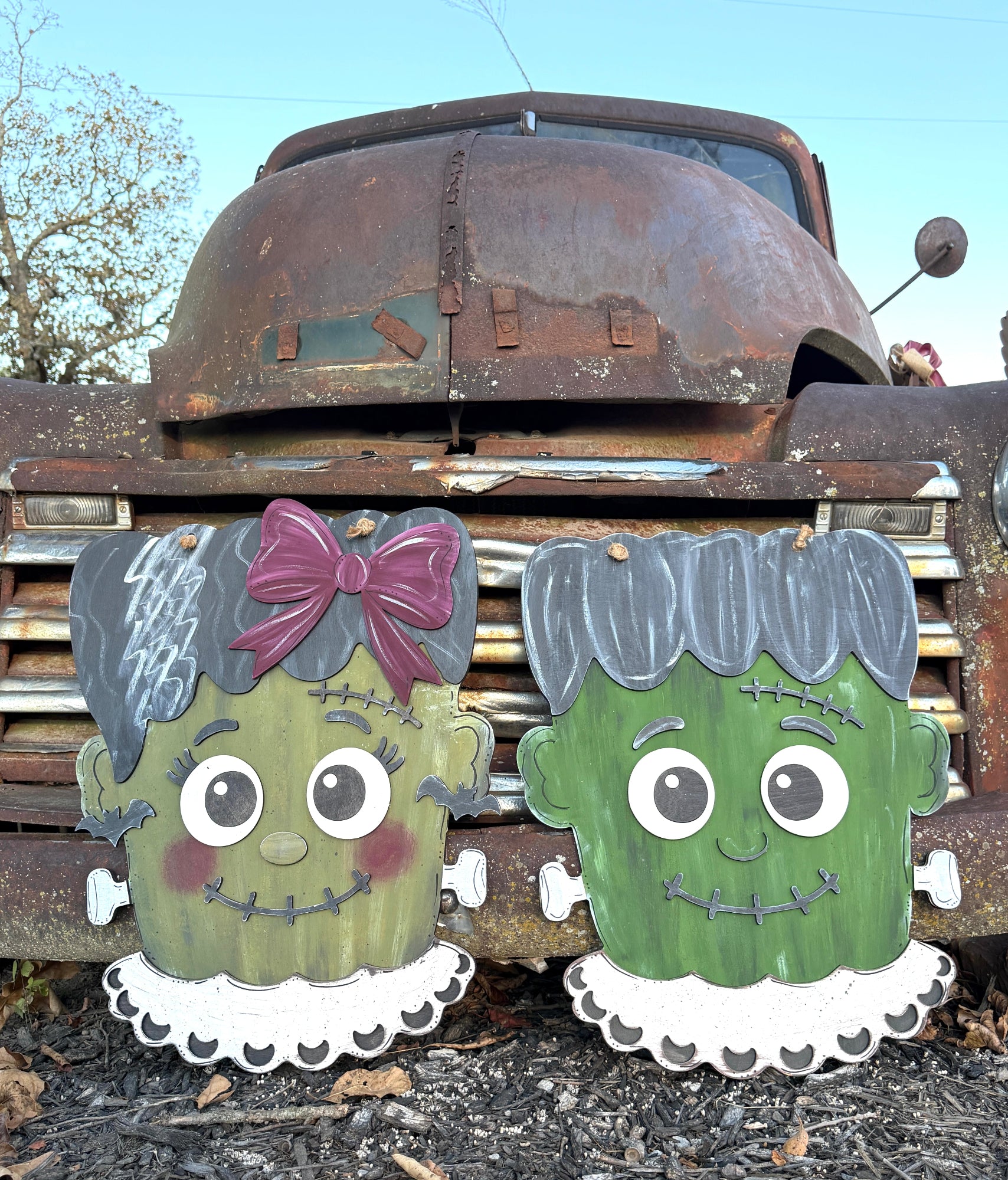 Two painted wooden Frankenstein-themed yard signs in front of a rusted vintage truck outdoors. One sign depicts a green female monster with a purple bow and white ruffled collar, and the other depicts a green male monster with stitched hair and a white ruffled collar.
