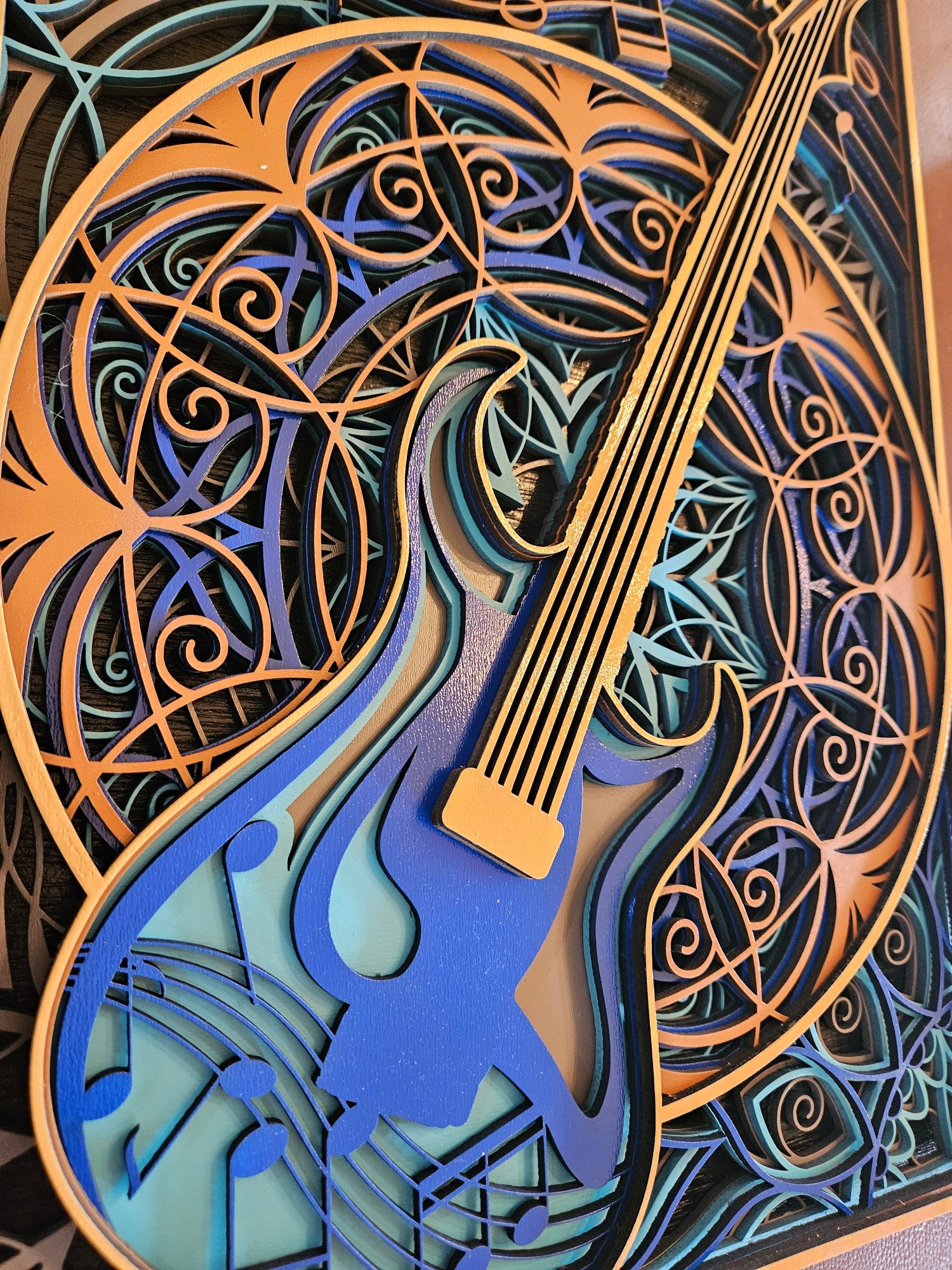 A close-up of a layered laser-cut wooden wall art piece shaped like an electric guitar, featuring blue, teal, and orange wood tones with intricate swirling patterns.