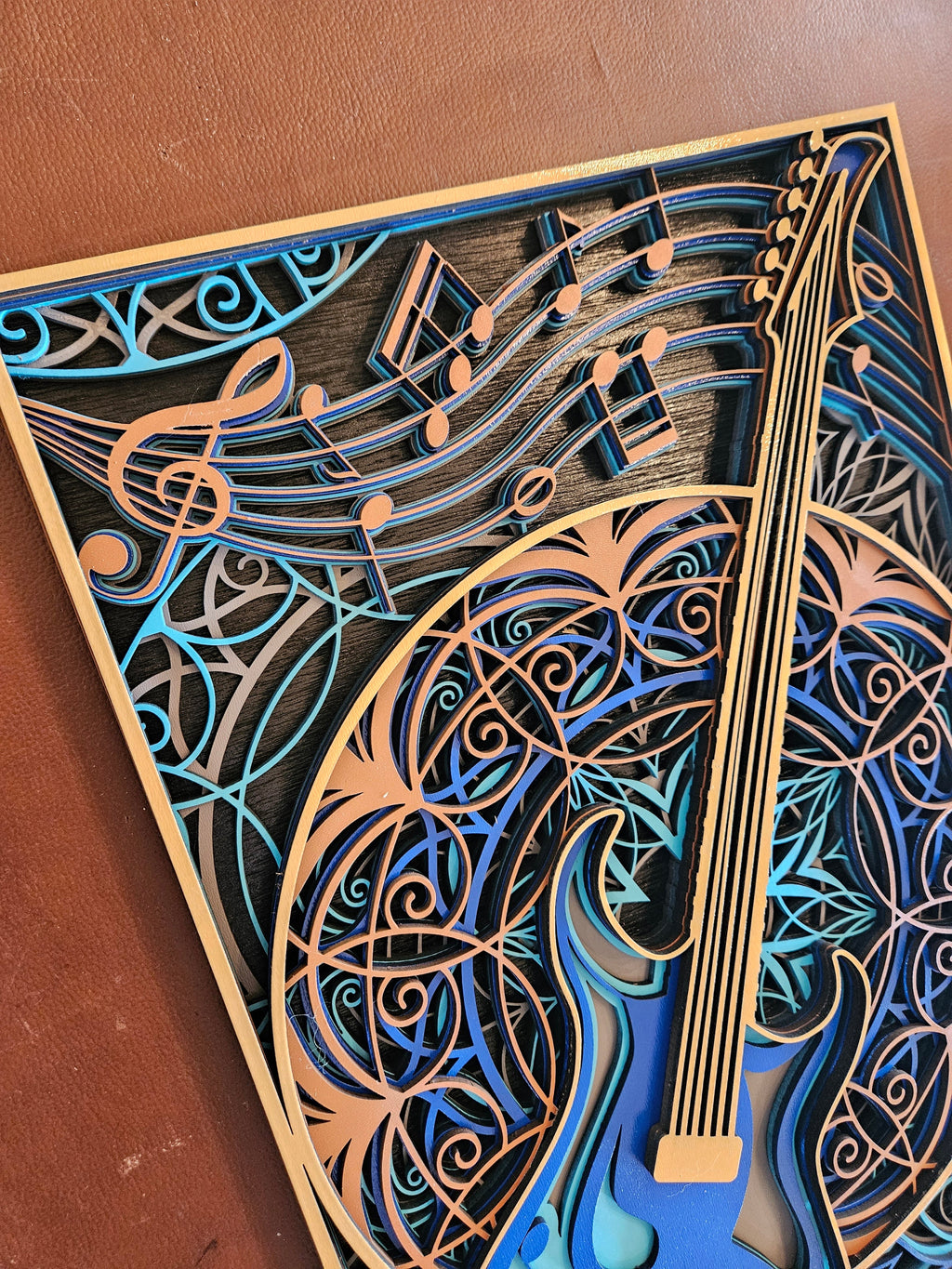 A close-up of a laser-cut wooden wall art piece featuring a stylized guitar and musical notes, layered in shades of blue, teal, and natural wood tones on a brown surface.