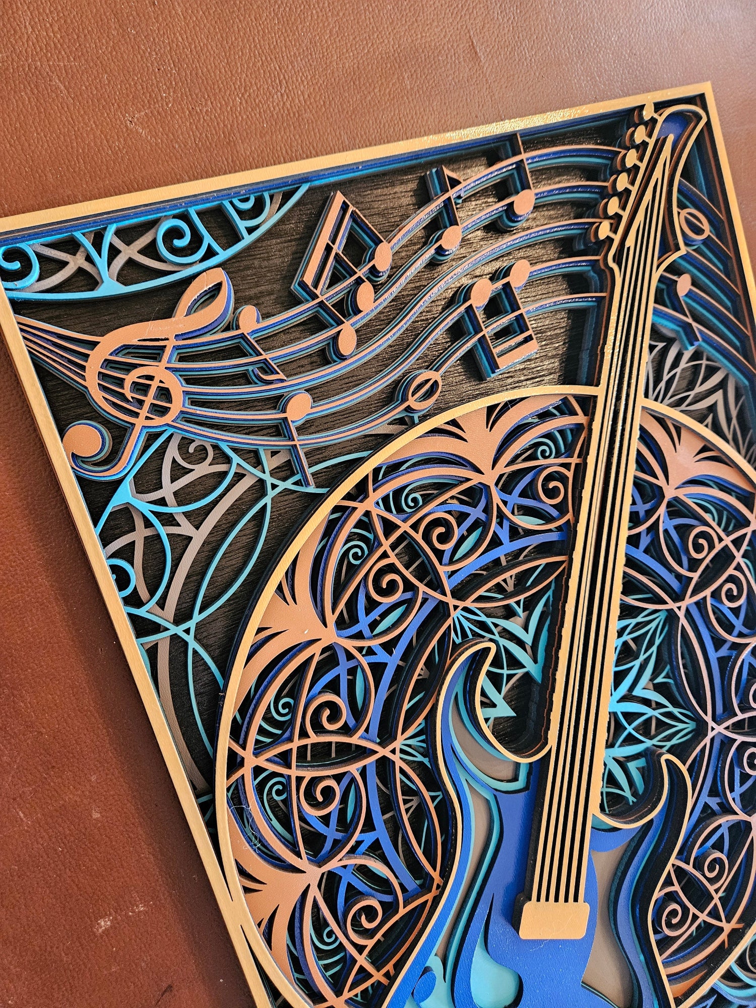 A close-up of a laser-cut wooden wall art piece featuring a stylized guitar and musical notes, layered in shades of blue, teal, and natural wood tones on a brown surface.