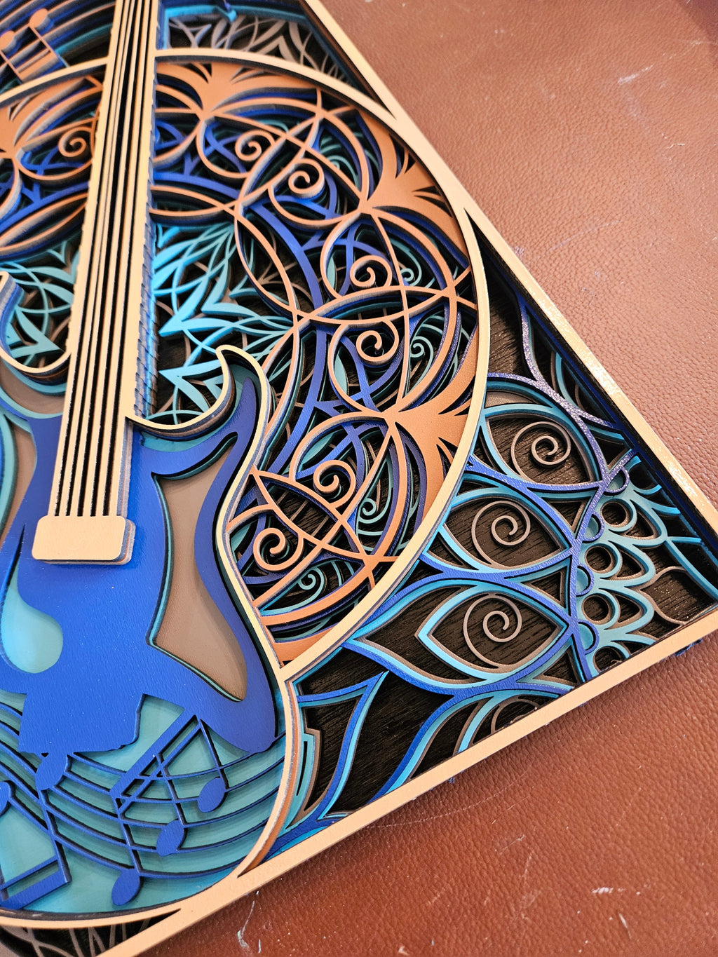 A close-up of a laser-cut wooden wall art piece featuring a blue electric guitar and swirling geometric patterns in layered wood tones, shown on a brown textured surface.