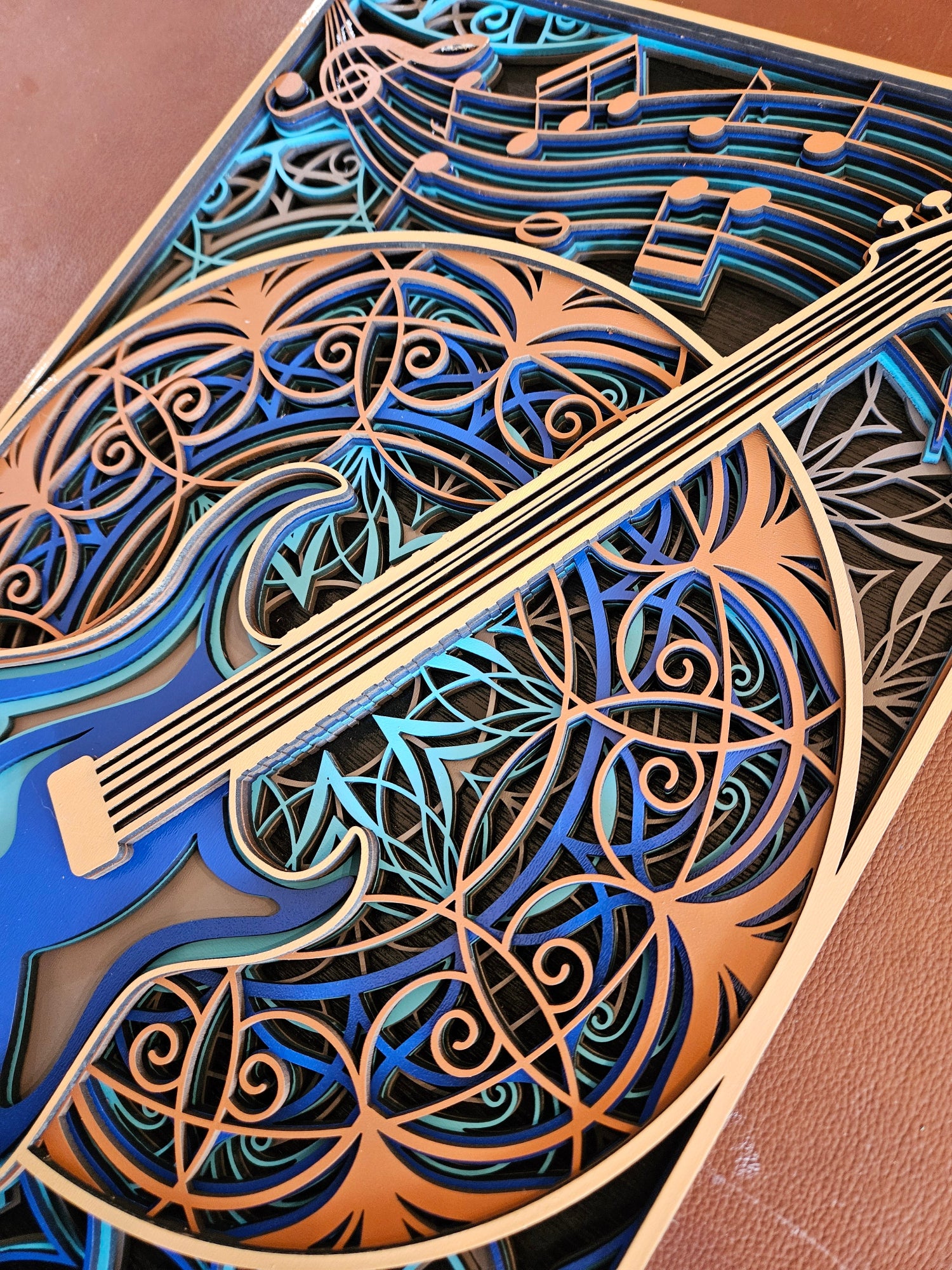 A close-up of a laser-cut wooden wall art piece featuring a violin and musical notes, with layered wood in natural tan, blue, and black tones.